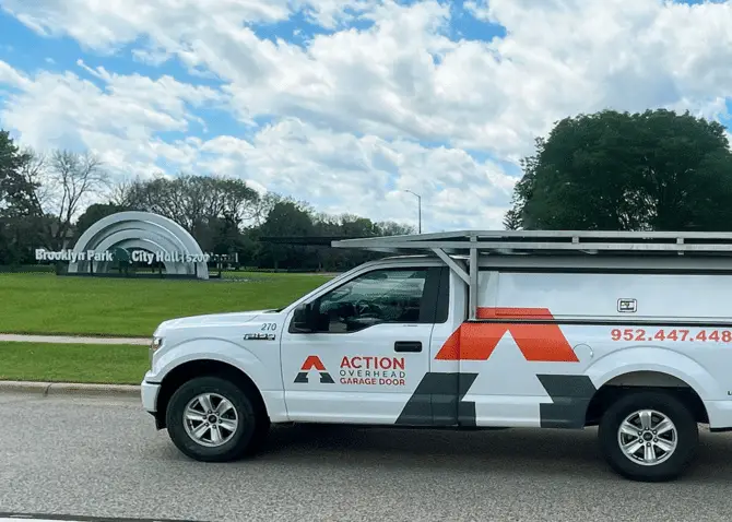 A white Action service truck is parked outside the Brooklyn Park city hall sculpture under a partly blue sky.
