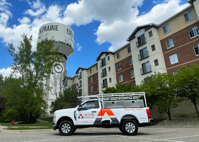 A white Action Overhead pick-up truck sits underneath the Eden Prairie water tow near a row condominiums. Landcaping trees in full bloom and clouds drift overhead across a deep blue sky..